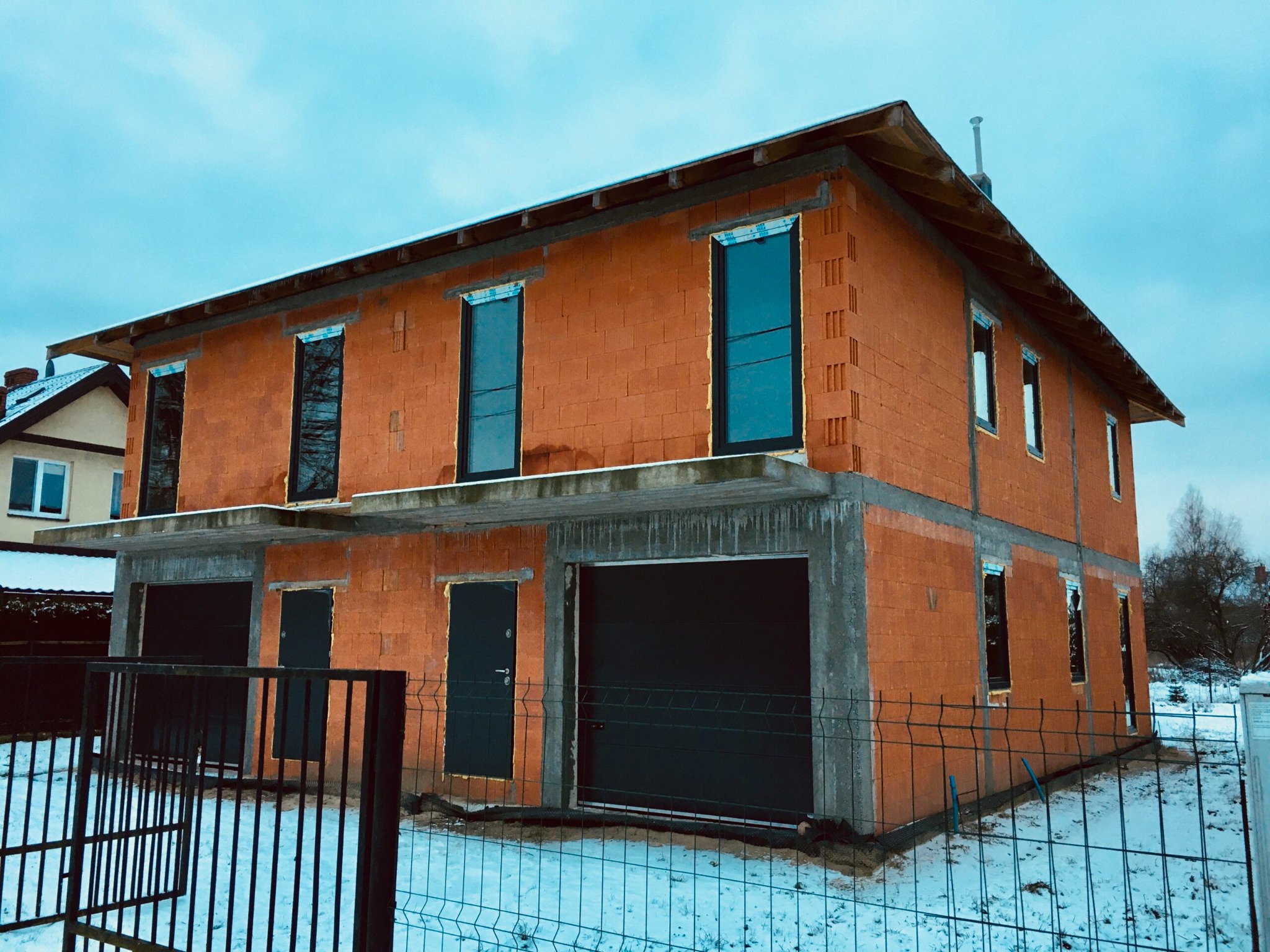 Dwukondygnacyjny dom w stanie surowym otwartym, z widocznymi murami z czerwonej cegły, wstępnie zamontowanymi oknami z czarnymi ramami i garażem, otoczony ogrodzeniem siatkowym w zimowej scenerii.