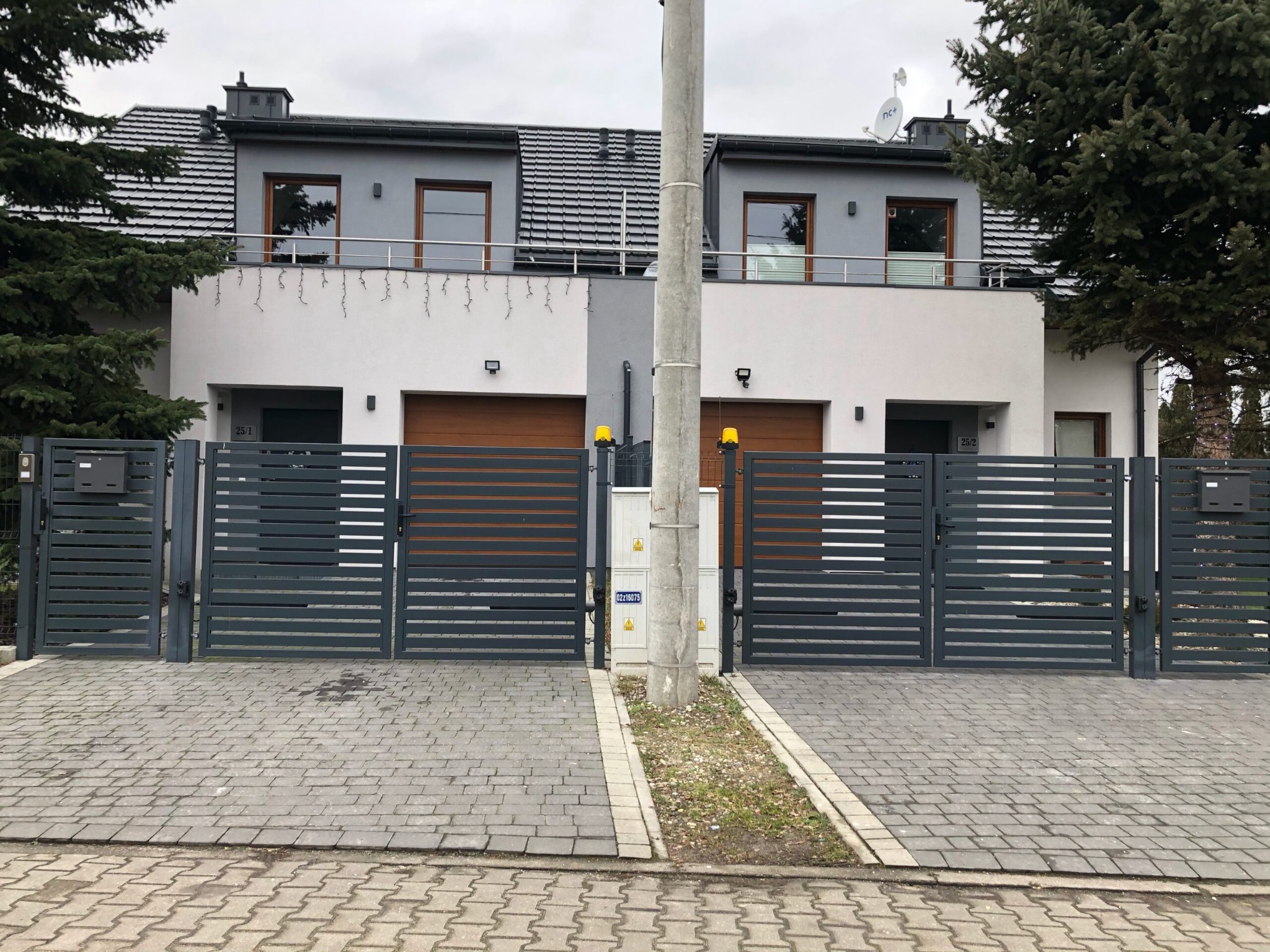 Dwa bliźniacze domy z szarymi elewacjami, drewnianymi bramami garażowymi i nowoczesnymi, metalowymi ogrodzeniami, oddzielone słupem energetycznym na tle pochmurnego nieba.