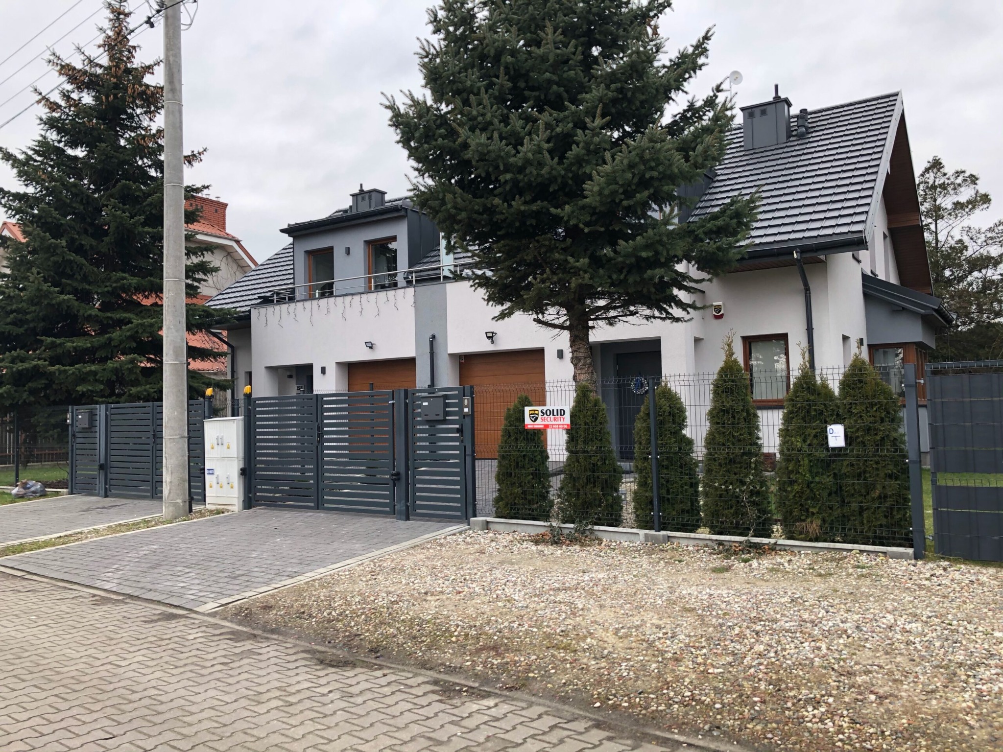 Nowoczesny dom jednorodzinny z szarą dachówką, ocieploną elewacją, grafitową bramą wjazdową i zadbanym otoczeniem z iglakami.