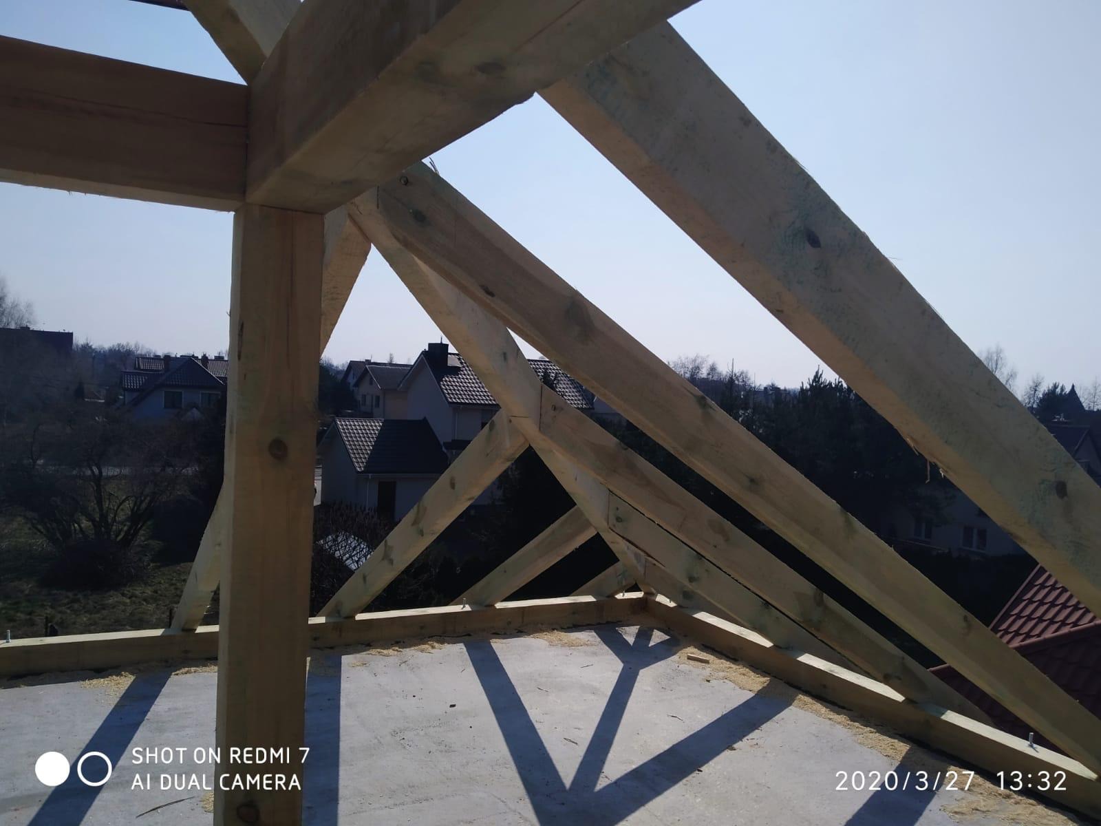 Wnętrze domu w trakcie budowy dachu, widoczne drewniane krokwie i słupy konstrukcyjne na tle niebieskiego nieba i zabudowań mieszkalnych.