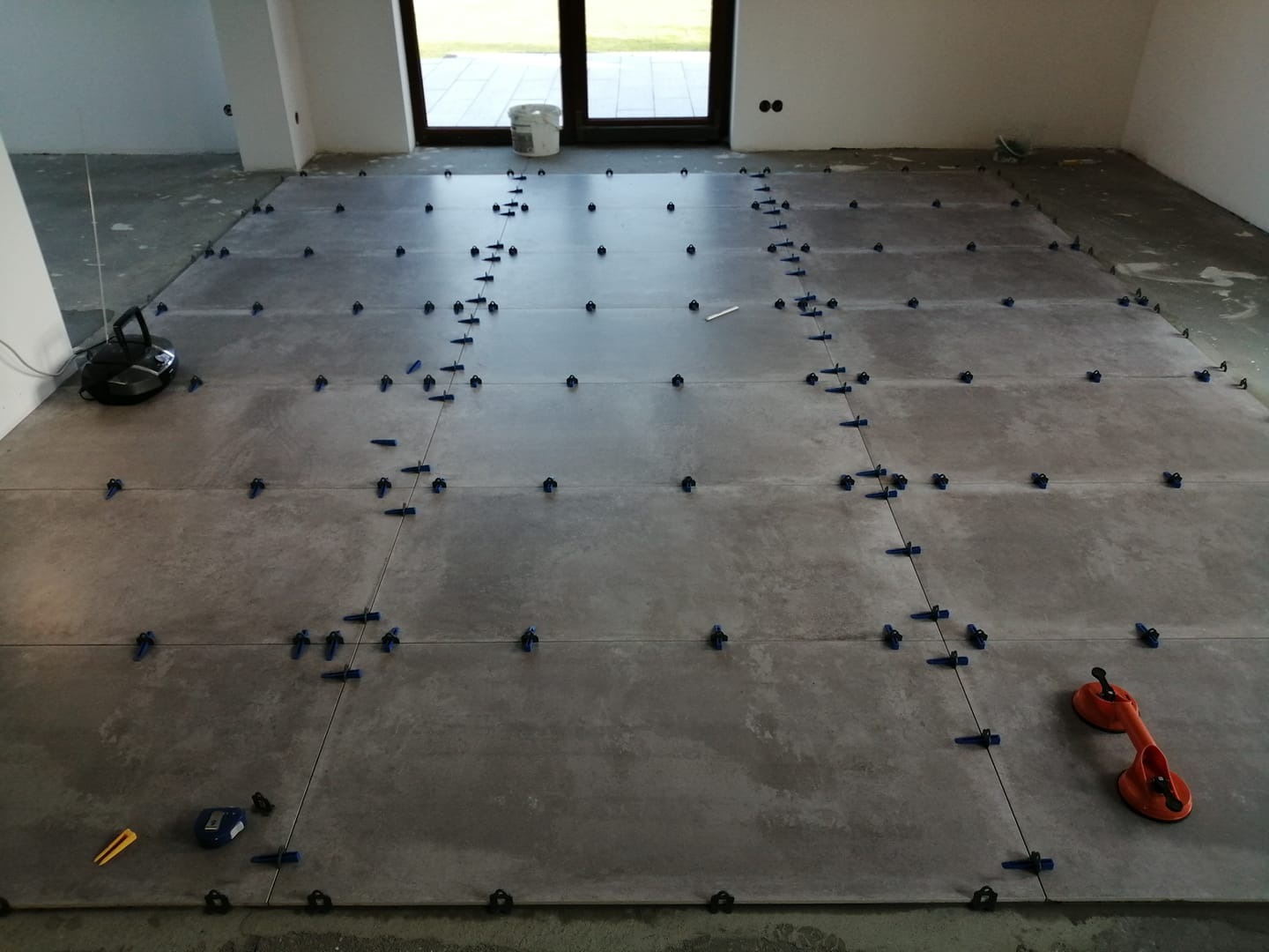 Układanie szarych płytek podłogowych w nowym budynku z użyciem niebieskich klipsów poziomujących i przyssawki do przenoszenia płytek.