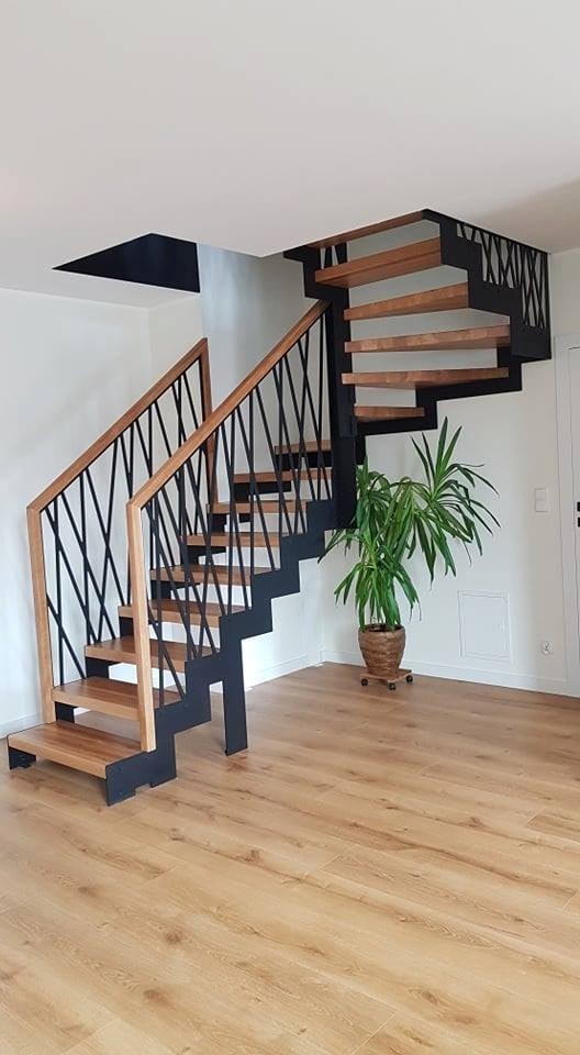 Nowoczesne schody drewniane z czarną metalową konstrukcją i oryginalną balustradą krzyżową, oświetlone naturalnym światłem, z rośliną doniczkową w tle na drewnianej podłodze.