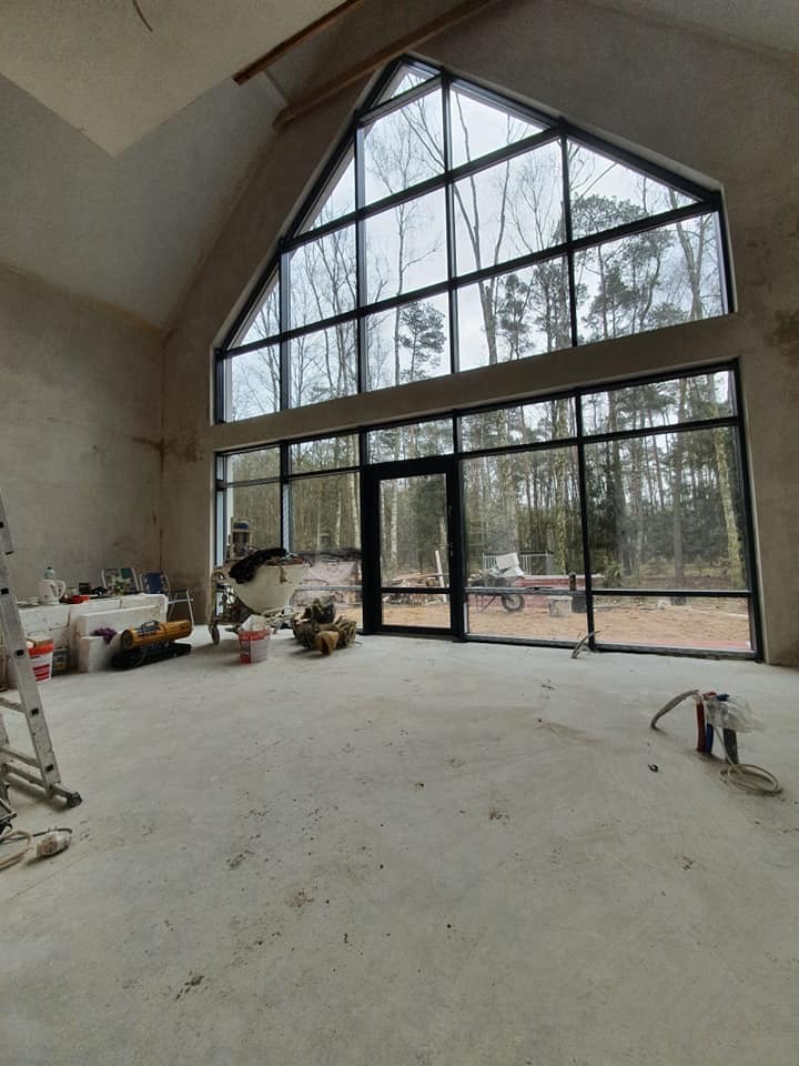 Surowe wnętrze z wysokim, trójkątnym oknem z czarną ramą, przez które widać las, oraz rozstawionymi narzędziami i materiałami budowlanymi, sugerujące etap prac wykończeniowych.