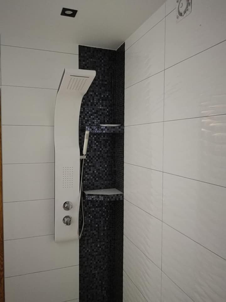 Nowoczesna kabina prysznicowa z panelem natryskowym i czarną mozaiką z półkami w wykończonej łazience, białe płytki z delikatną fakturą na ścianach.