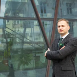 Młody mężczyzna w tweedowym garniturze z zielonym krawatem i poszetką, stoi na tle nowoczesnego budynku ze szklaną fasadą.