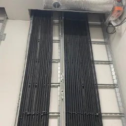 Rząd czarnych kabli elektrycznych ułożonych w metalowej drabince przymocowanej do ściany, widoczny przewód wentylacyjny w srebrnej folii oraz czerwona rurka pod sufitem.