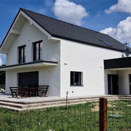 OSKOSTUDIO - Nowoczesny dom jednorodzinny z białymi ścianami i ciemną stolarką okienną, widok z zewnątrz w słoneczny dzień. Na tarasie drewniany stół i ławki.