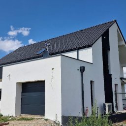 OSKOSTUDIO - Nowoczesny dom jednorodzinny z garażem, biała elewacja z czarnymi akcentami, dachówka w kolorze grafitu, widok z boku na tle błękitnego nieba z chmurami.