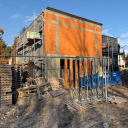 OSKOSTUDIO - Budowa domu jednorodzinnego w trakcie realizacji: ściany z czerwonej cegły i szare ocieplenie, rusztowania, palety i ogrodzenie budowlane na pierwszym planie.