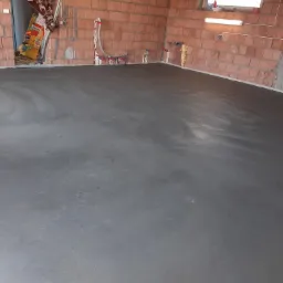 Świeżo wylana, gładka posadzka w surowym wnętrzu z czerwonej cegły, widoczne instalacje wodne i elektryczne, okno w górnej części ściany.