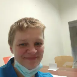 Uśmiechnięta kobieta w niebieskim uniformie i maseczce pod brodą, za biurkiem z dokumentami i przegrodą z pleksi.