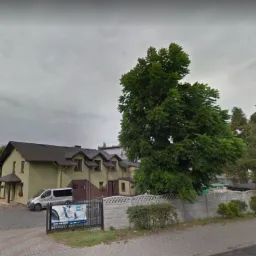 Zewnętrzny widok budynku warsztatu samochodowego z zaparkowanym białym vanem, otoczonego zielenią i ogrodzeniem, z reklamą na ogrodzeniu.