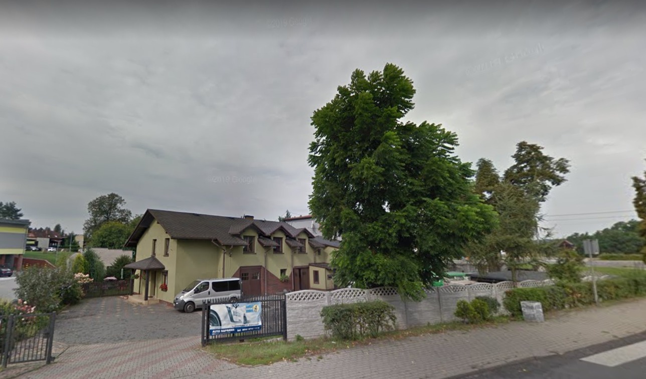 Zewnętrzny widok budynku warsztatu samochodowego z zaparkowanym białym vanem, otoczonego zielenią i ogrodzeniem, z reklamą na ogrodzeniu.