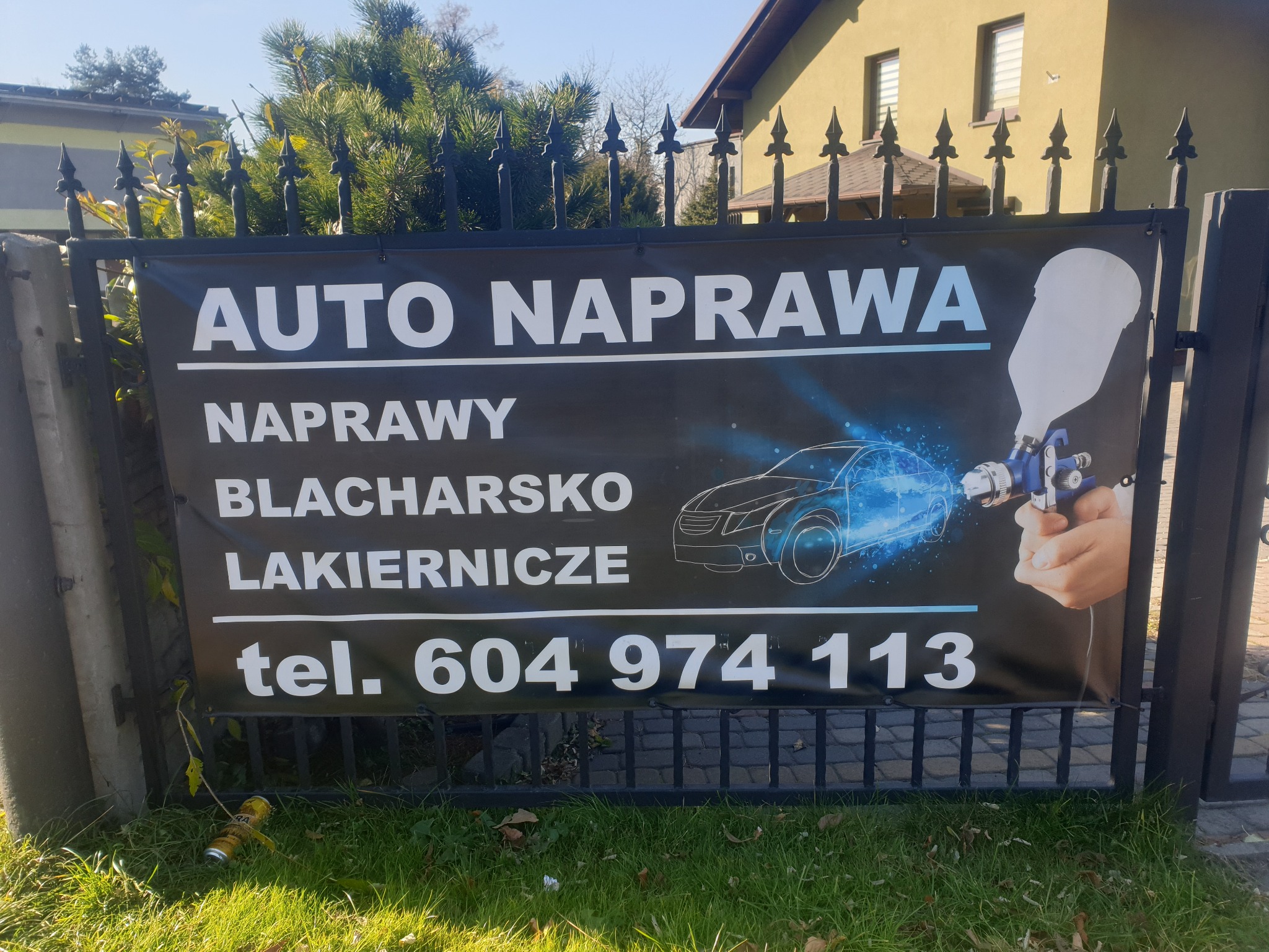 Baner reklamowy 'Auto Naprawa' zawieszony na metalowym ogrodzeniu przed budynkiem; w tle widoczna trawa i puszka po napoju.