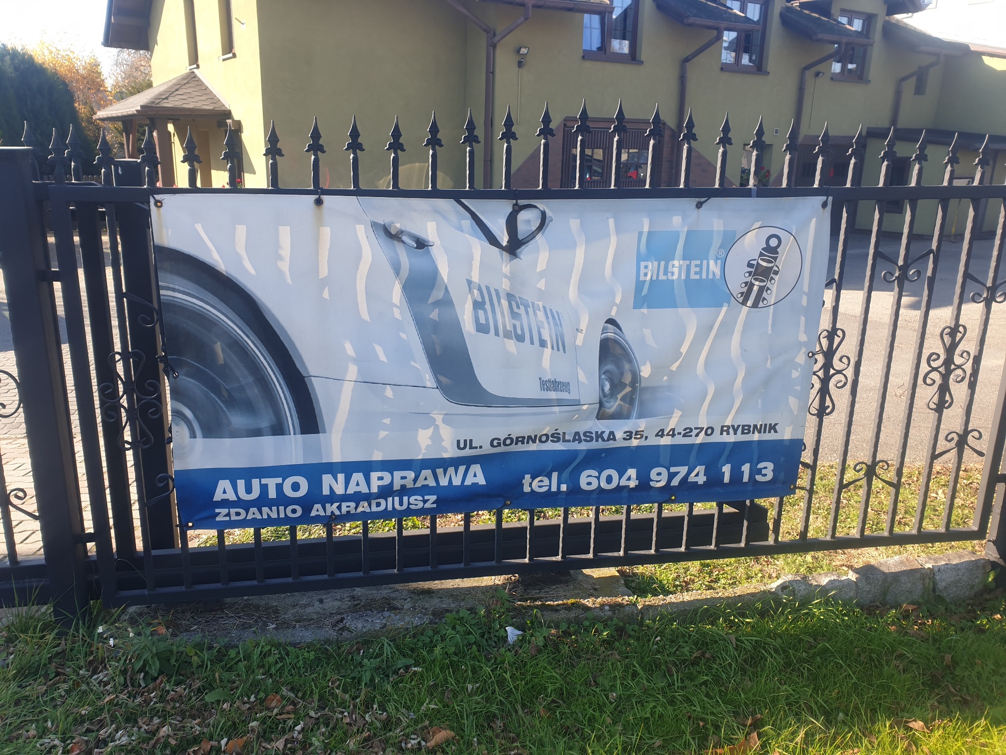 Reklama warsztatu samochodowego 'Auto Naprawa Zdanio Akradiusz' z logo Bilstein, umieszczona na metalowym ogrodzeniu przed budynkiem.