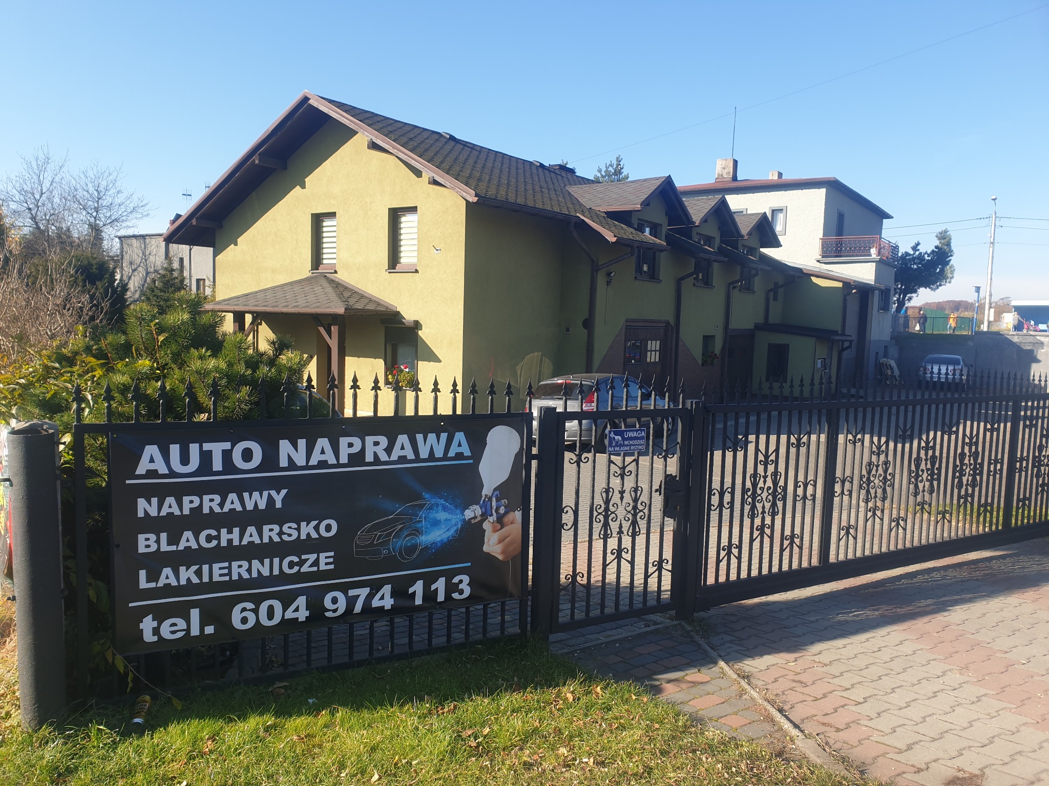 Tablica reklamowa warsztatu samochodowego z numerem telefonu, umieszczona na tle budynku i ogrodzenia.