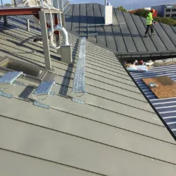 Szary dach z blachy trapezowej z zamontowanymi elementami komunikacji dachowej oraz pracownikiem w odblaskowej kamizelce, widoczny fragment instalacji wentylacyjnej i komin.