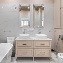 Elegancka łazienka z dwoma umywalkami nablatowymi, lustrami w prostokątnych ramach, wanną wolnostojącą i zabudową meblową w odcieniach beżu i bieli z marmurowymi płytkami na ścianach i podłodze.