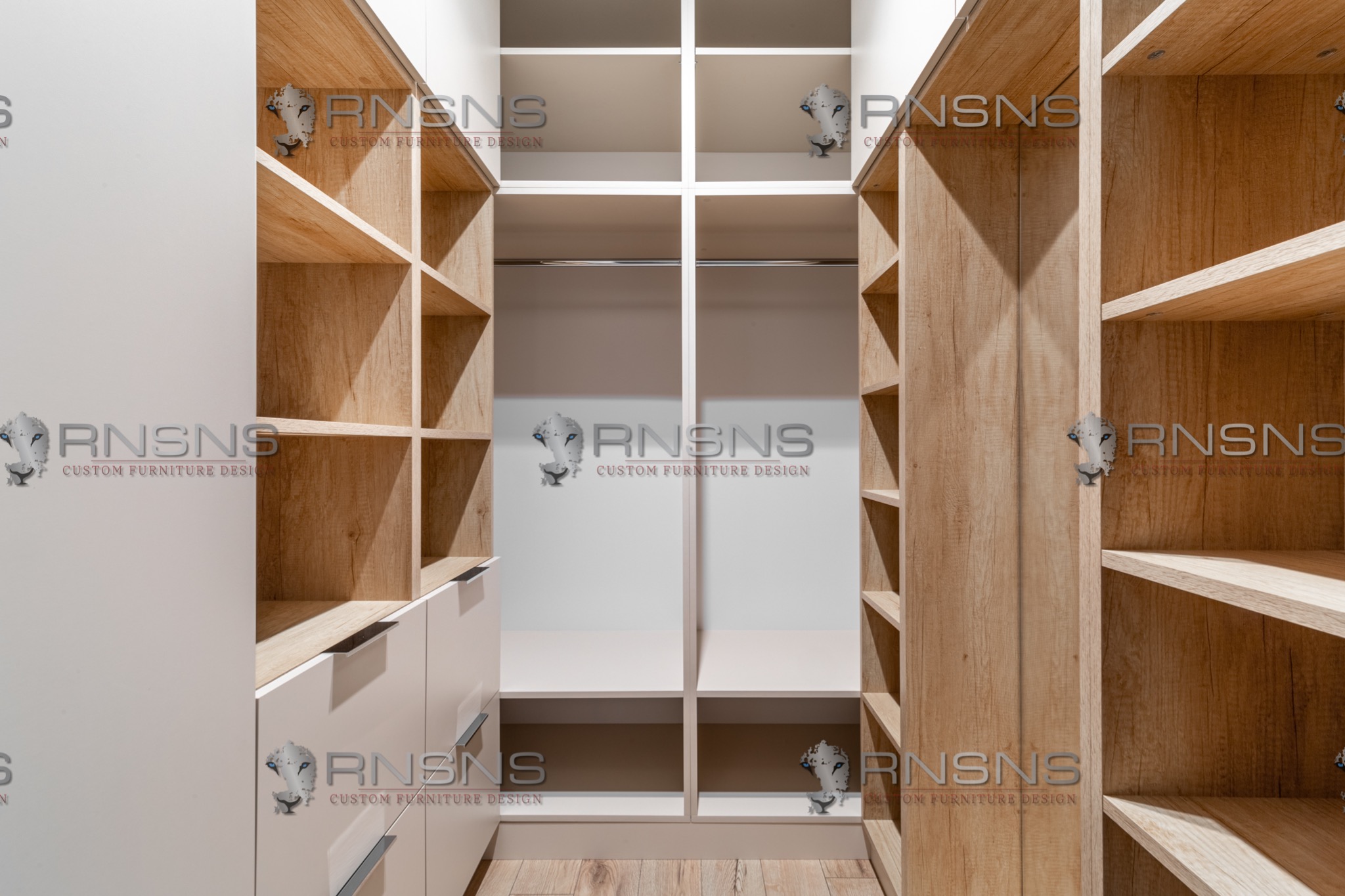 Wnętrze przestronnej szafy wnękowej z półkami z jasnego drewna i białymi elementami, widok z perspektywy wejścia, z widocznym logo firmy na elementach szafy.