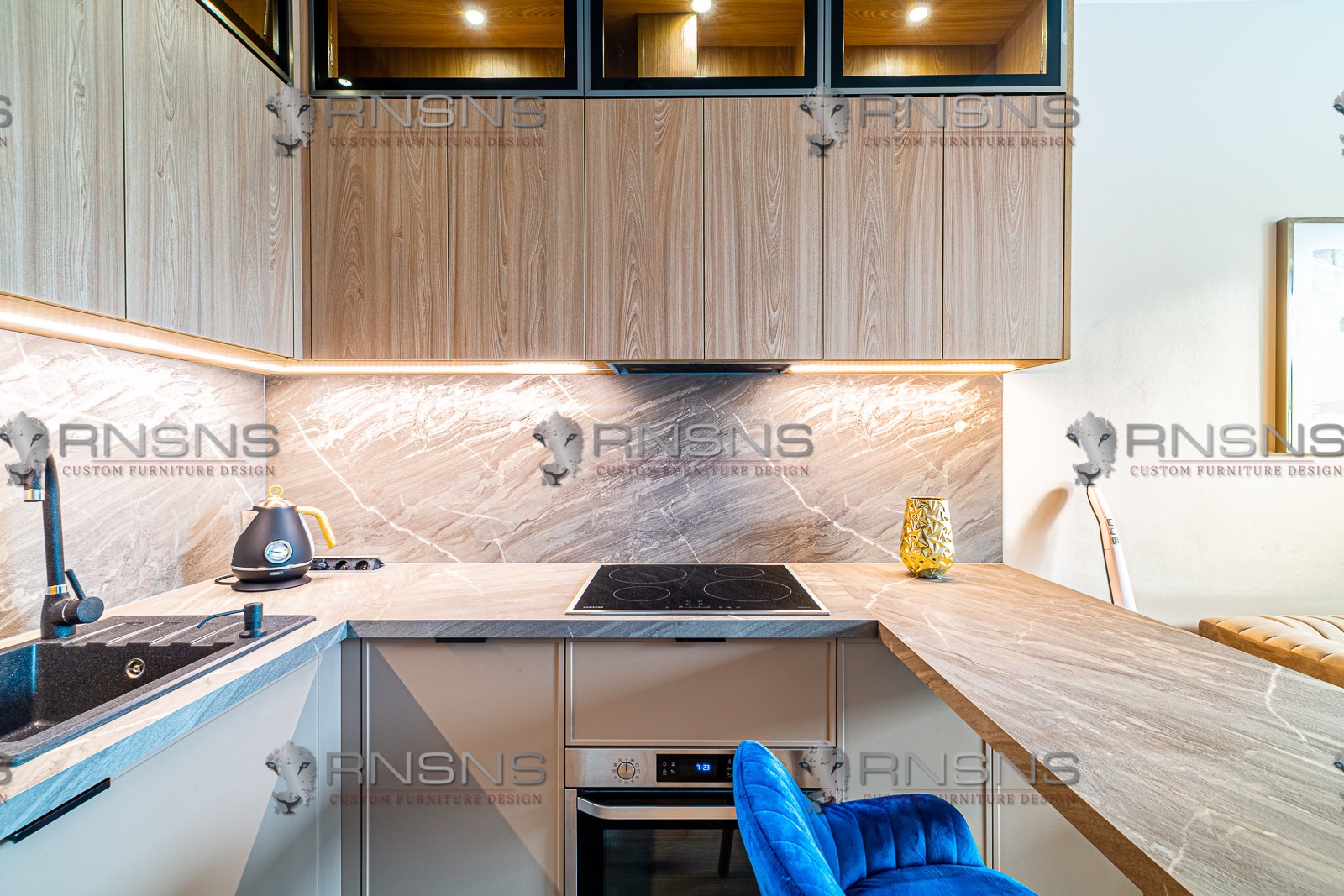Nowoczesna kuchnia z drewnianymi szafkami, marmurowym blatem, czarnym zlewem i płytą indukcyjną. Niebieski krzesło barowe dodaje koloru. Oświetlenie LED pod szafkami górnymi.