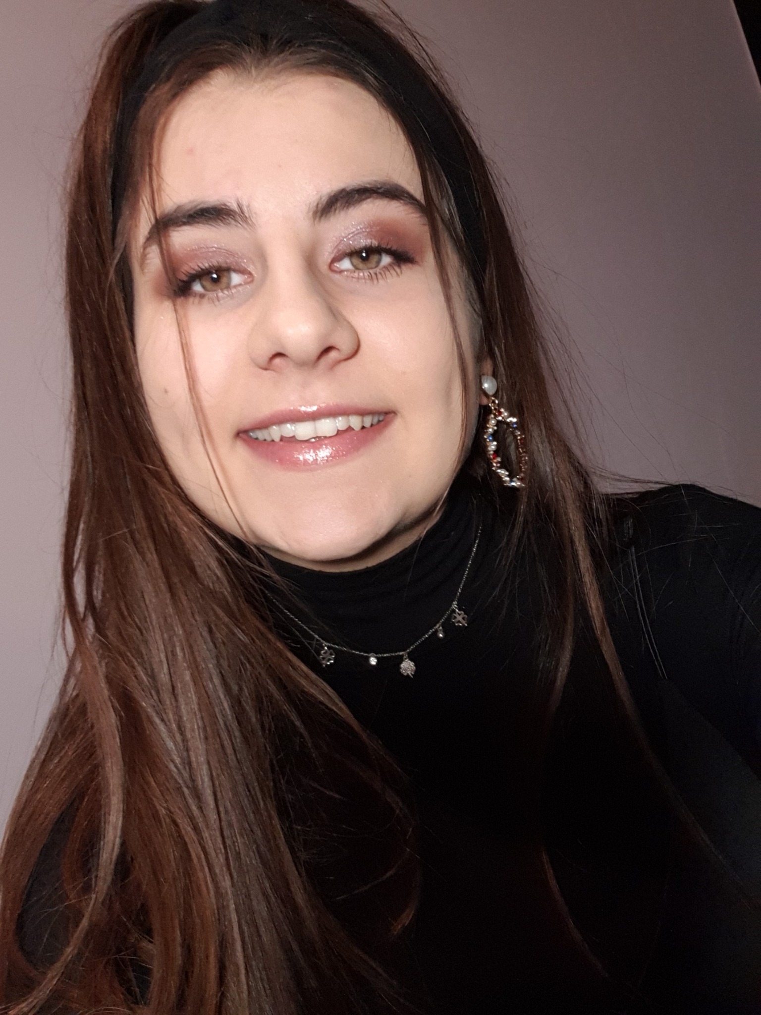 Portret uśmiechniętej brunetki w czarnym golfie, z delikatnym makijażem oczu i błyszczącymi ustami, noszącej ozdobne kolczyki i srebrny naszyjnik.