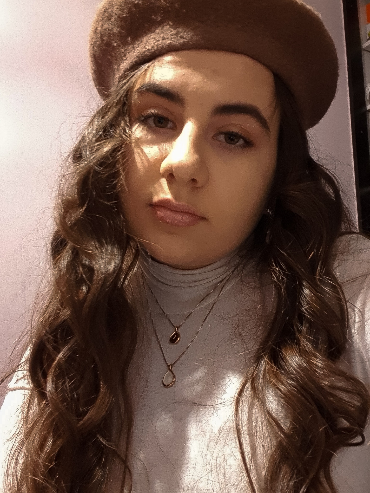 Portret młodej kobiety z brązowymi, kręconymi włosami, w beżowym berecie i białym golfie, z dwoma naszyjnikami, na tle jasnofioletowej ściany.