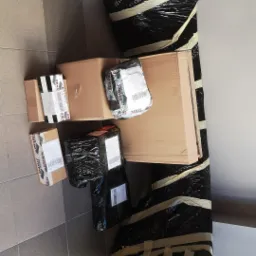 Zapakowane paczki różnej wielkości, w tym duży mebel owinięty czarną folią i taśmą, leżące na podłodze z płytek.