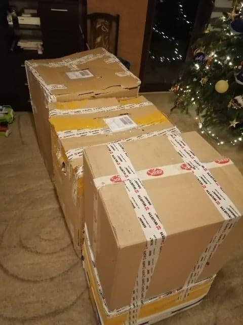 Stos kartonowych pudeł oklejonych taśmą transportową z logo Wurth i Sante, ustawionych na podłodze w pomieszczeniu z choinką w tle.