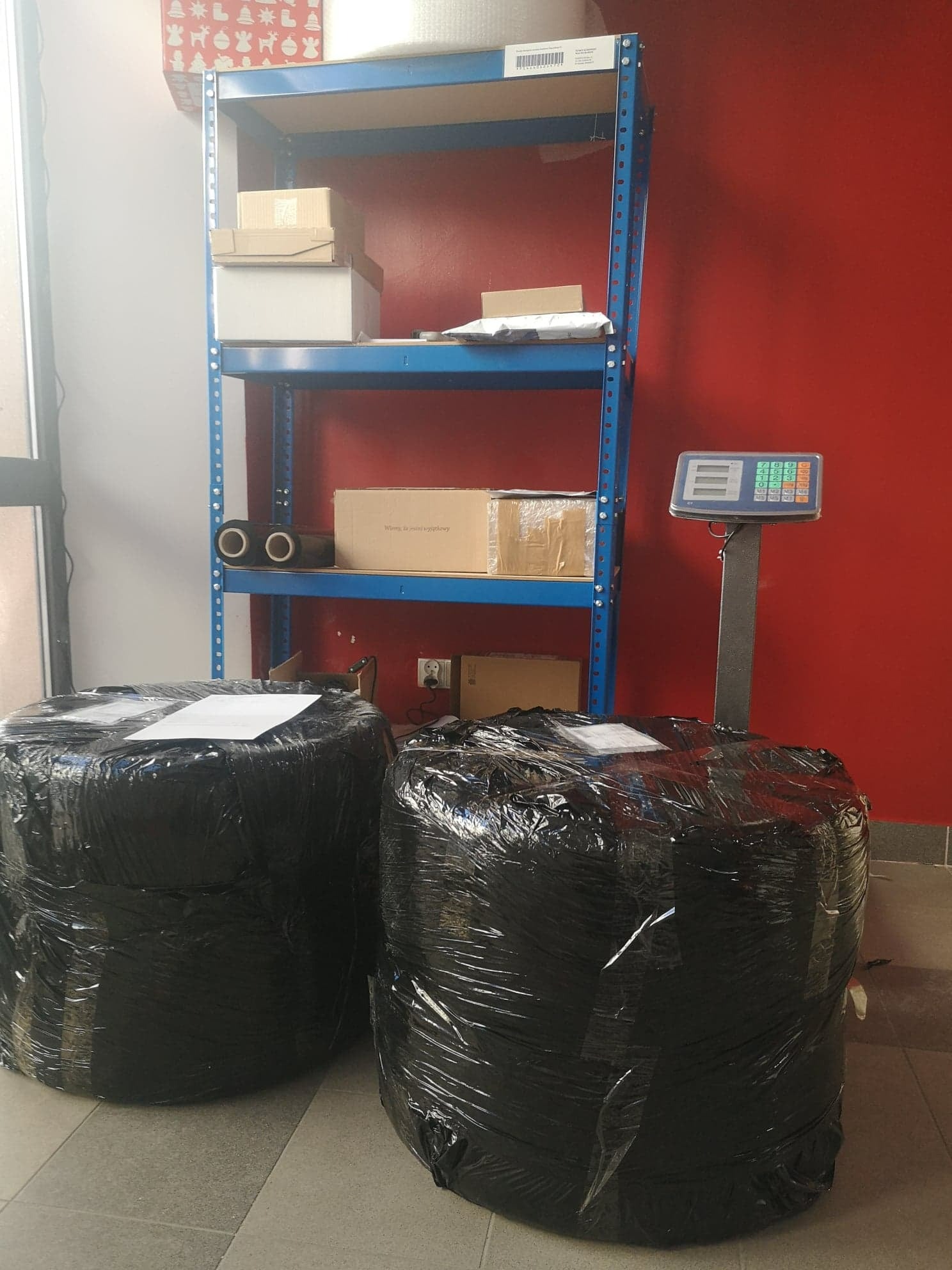 Dwie duże paczki owinięte czarną folią stretch, stojące obok wagi elektronicznej i metalowego regału z kartonami w tle.