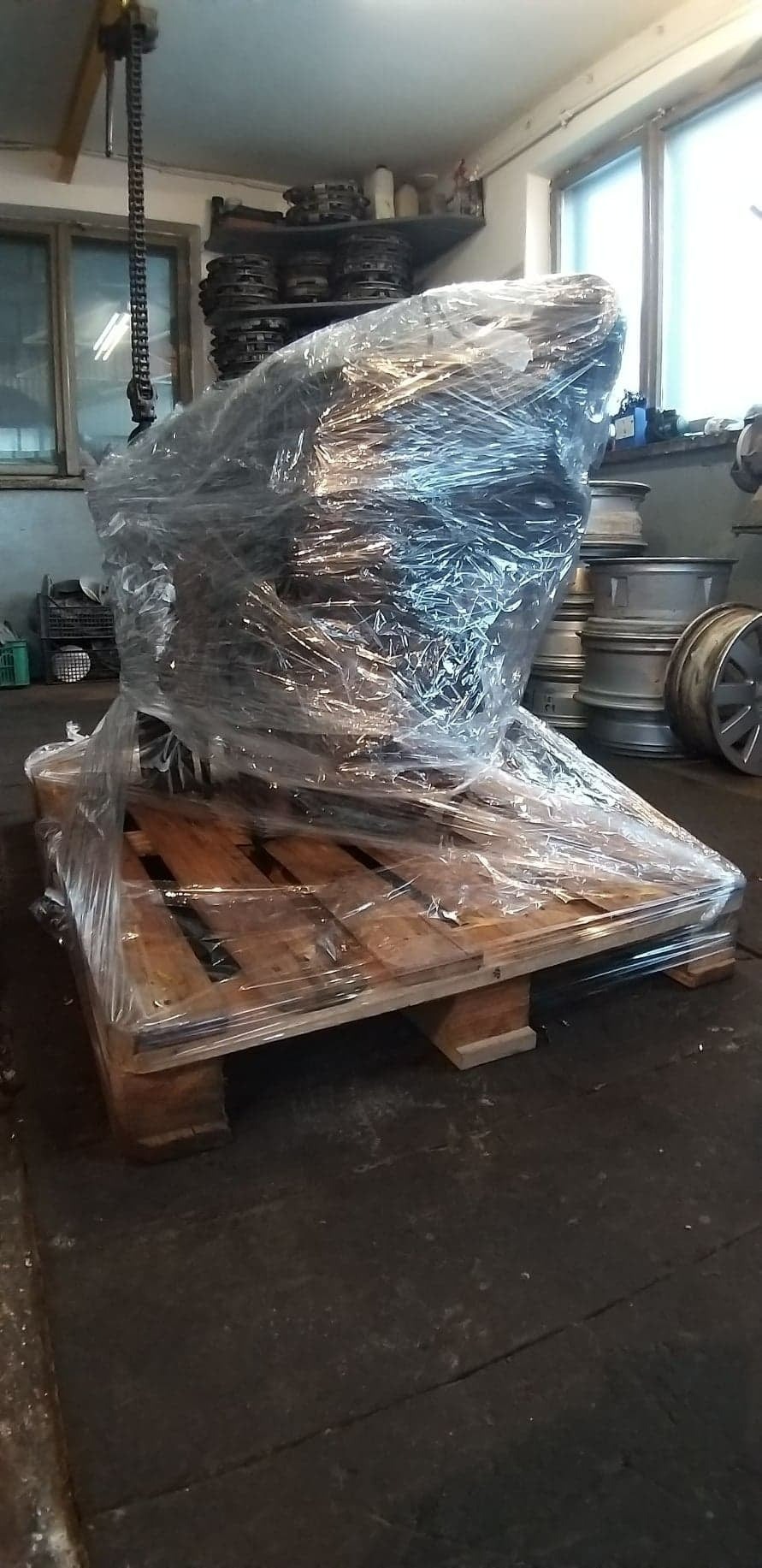 Silnik samochodowy zapakowany w folię na drewnianej palecie w warsztacie, widoczny wciągnik łańcuchowy i felgi samochodowe w tle.