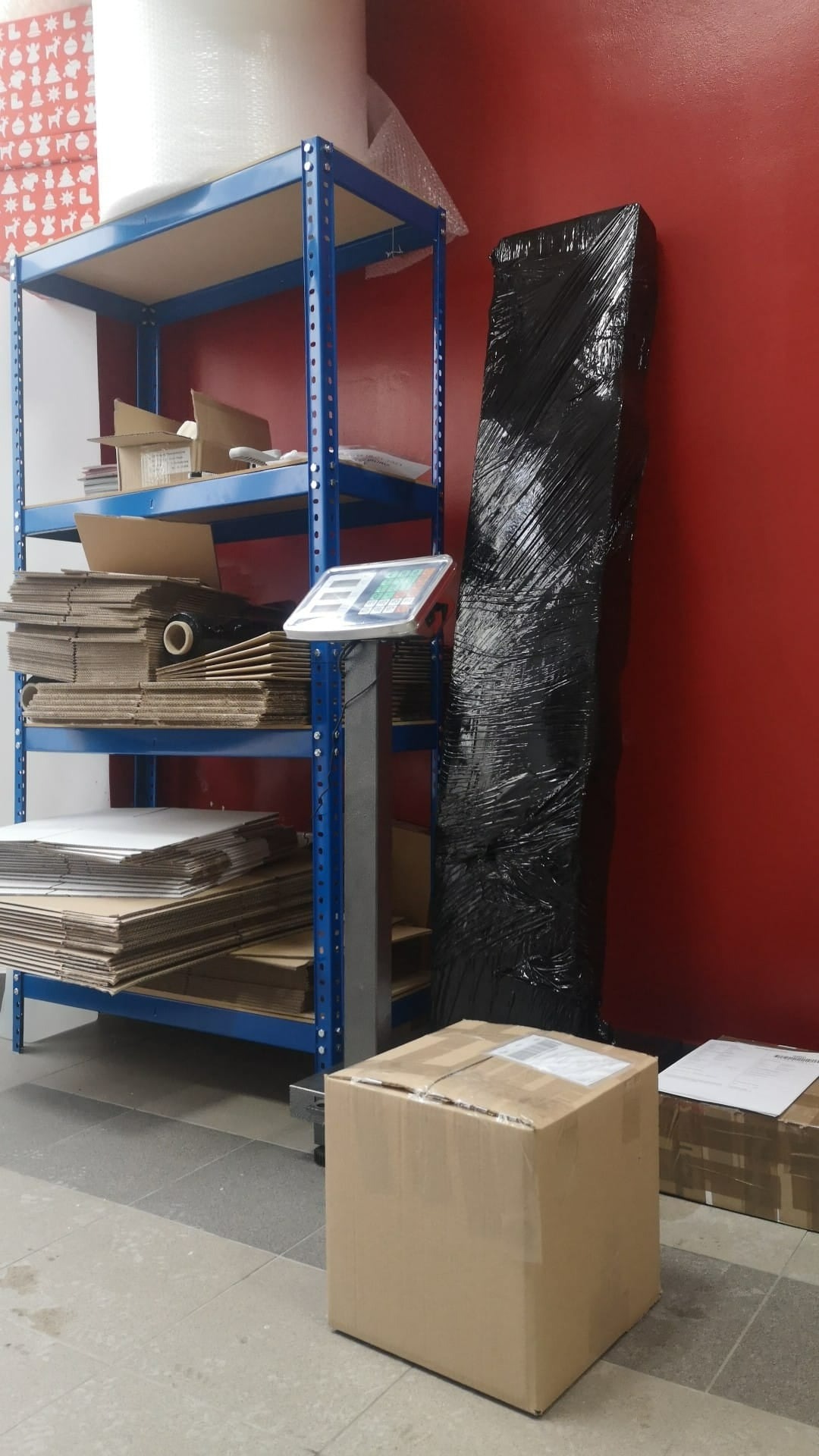 Stojący zapakowany w czarną folię przedmiot obok regału z kartonami i wagą, na podłodze kartonowe paczki.