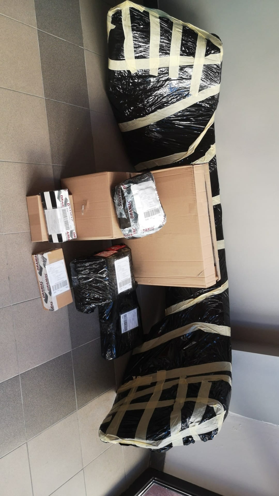 Zapakowane paczki różnej wielkości, w tym duży mebel owinięty czarną folią i taśmą, leżące na podłodze z płytek.