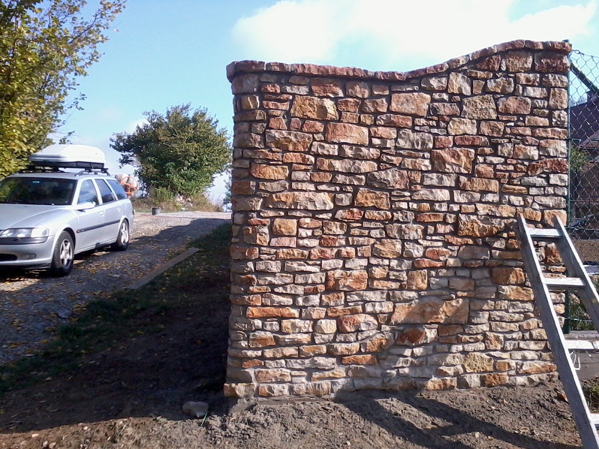 Kamienny murek z nieregularnych bloków piaskowca, ukończony z falistą górną krawędzią, z drabiną opartą o jego bok, na tle podjazdu z zaparkowanym srebrnym kombi z bagażnikiem dachowym.