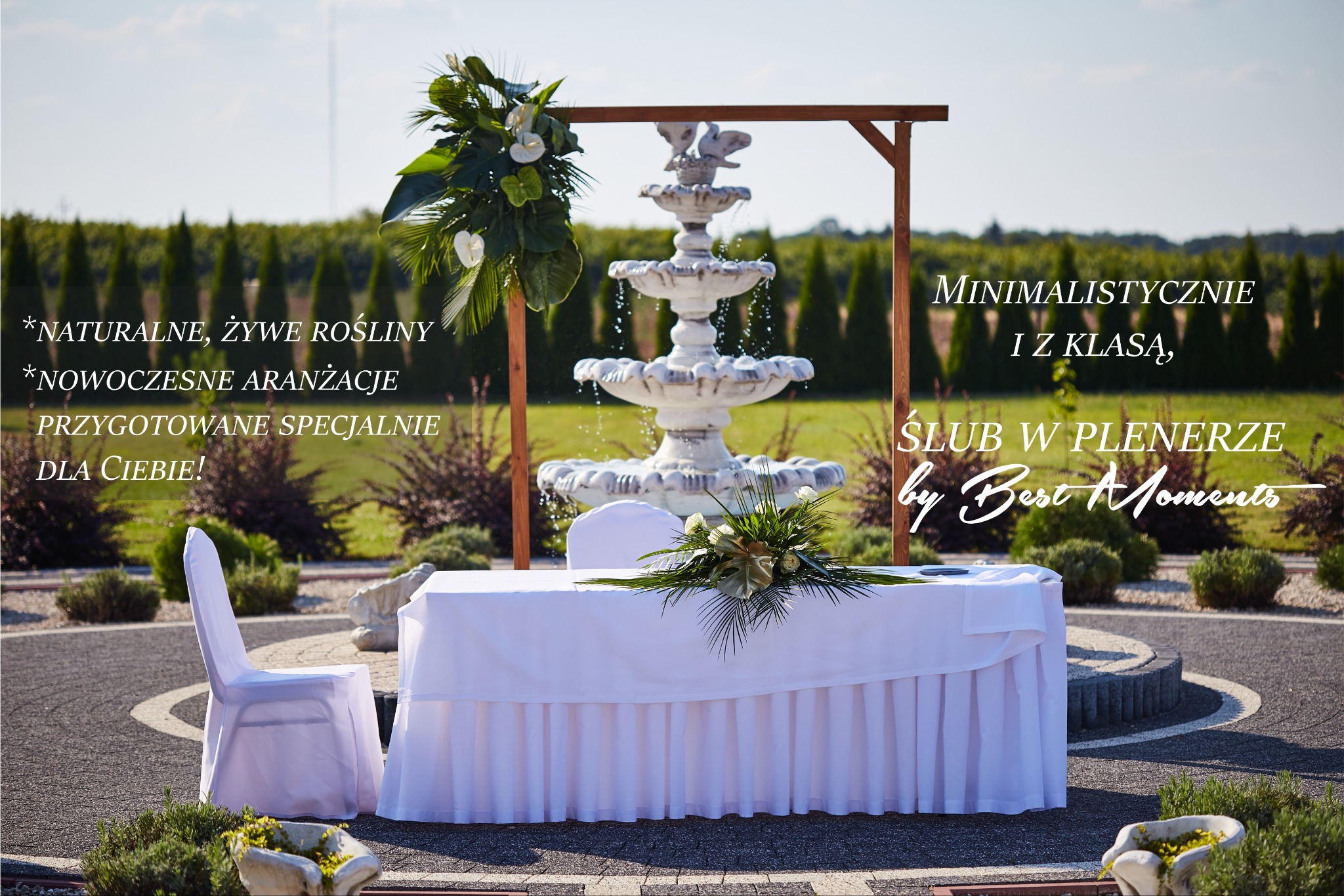 Dekoracja ślubna plenerowa: biały stół z falbaną i kwiatami, fontanna w tle, krzesło w pokrowcu, aranżacja roślinna na drewnianej konstrukcji.
