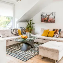 Jasny salon w skandynawskim stylu z narożną sofą, szklanym stolikiem kawowym na wzorzystym dywanie i dekoracyjnymi poduszkami z motywami zwierzęcymi, obraz z jeleniem na ścianie, duże okno z roletami.