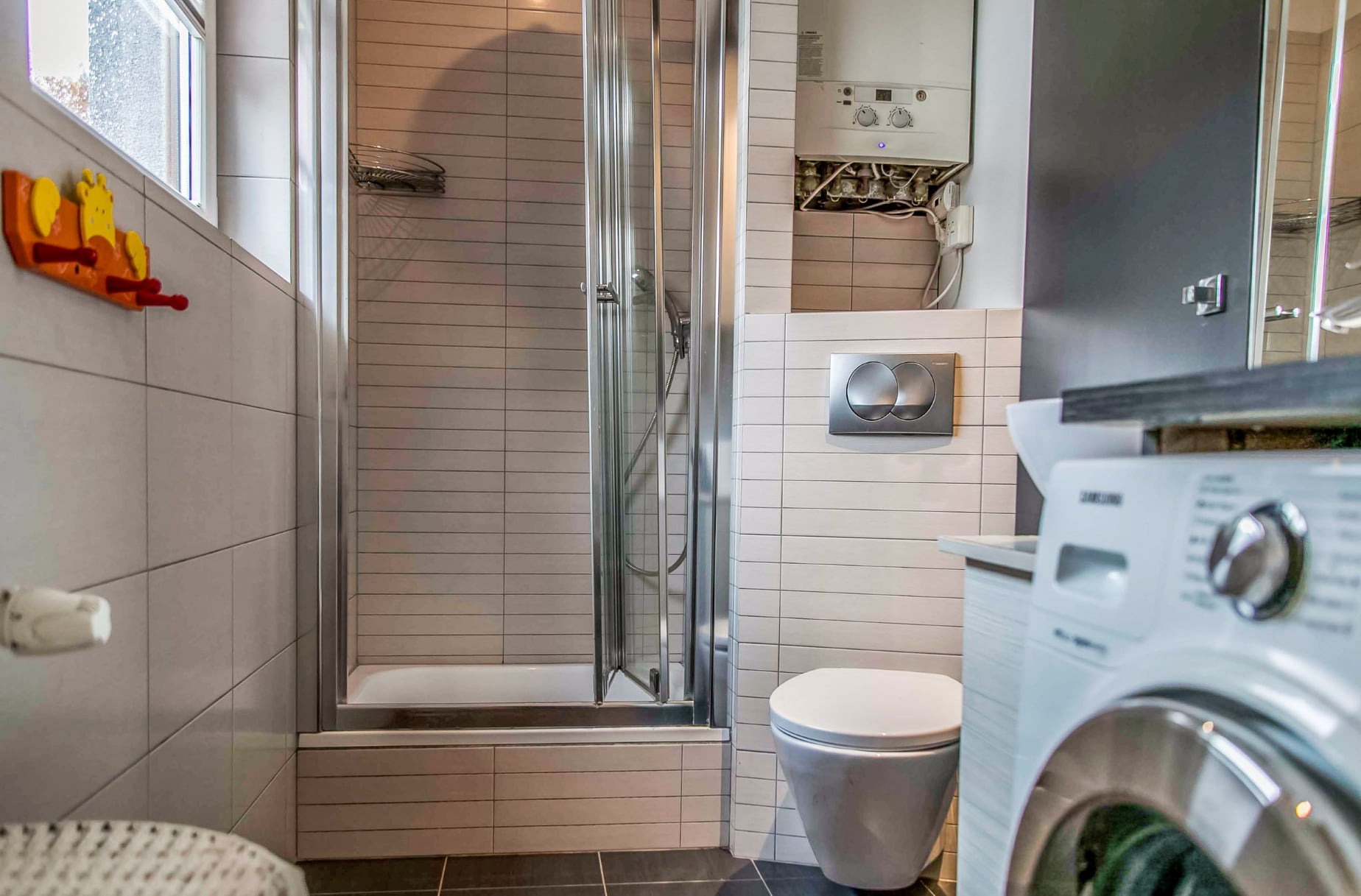 Wykończona łazienka w szaro-białej kolorystyce z kabiną prysznicową, podwieszaną toaletą, pralką Samsung i wiszącym piecem gazowym z widocznymi instalacjami.