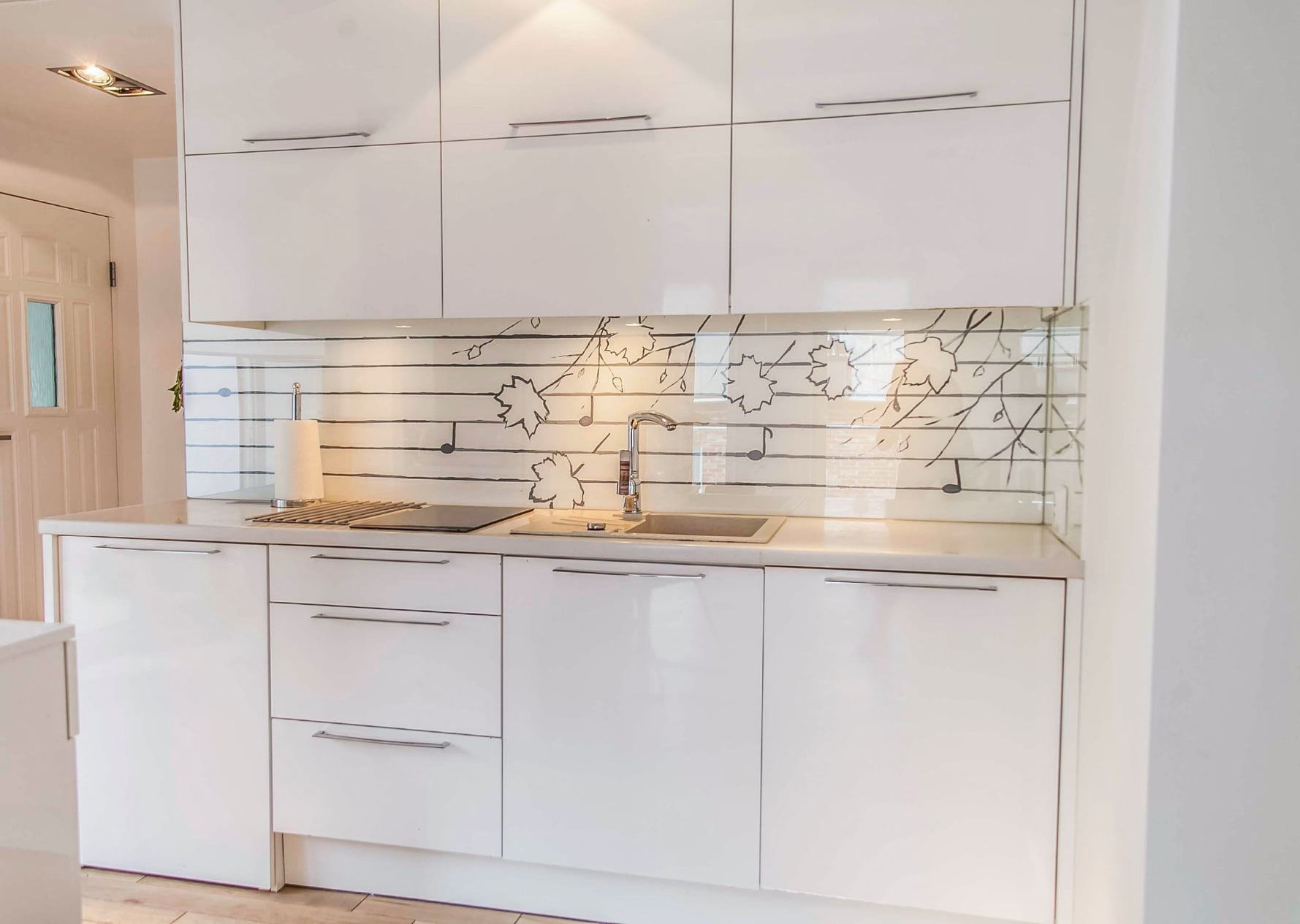 Biała, minimalistyczna kuchnia w Gdyni z błyszczącymi frontami szafek i szklanym panelem nad blatem z motywem liści i nut.
