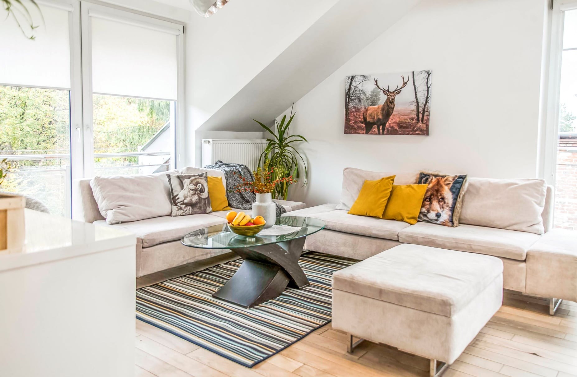 Jasny salon w skandynawskim stylu z narożną sofą, szklanym stolikiem kawowym na wzorzystym dywanie i dekoracyjnymi poduszkami z motywami zwierzęcymi, obraz z jeleniem na ścianie, duże okno z roletami.