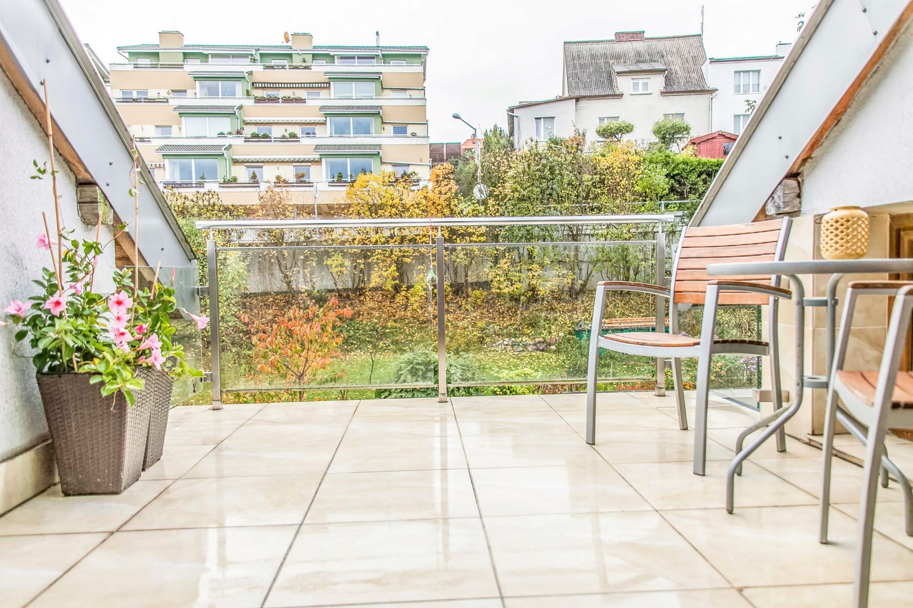 Błyszczący, beżowy taras z płytkami, szklana balustrada, drewniane krzesła i stolik, donice z różowymi kwiatami, widok na jesienny ogród i budynki.