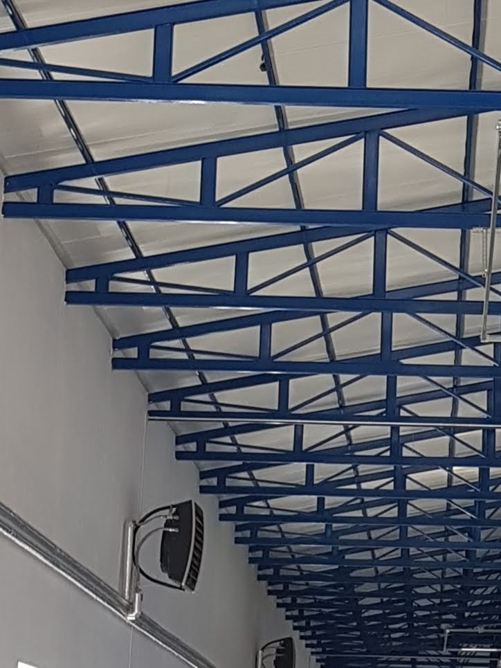 Wnętrze hali z widoczną konstrukcją dachu z niebieskich, stalowych belek kratowych i białym pokryciem, oświetlenie zamontowane na ścianie.
