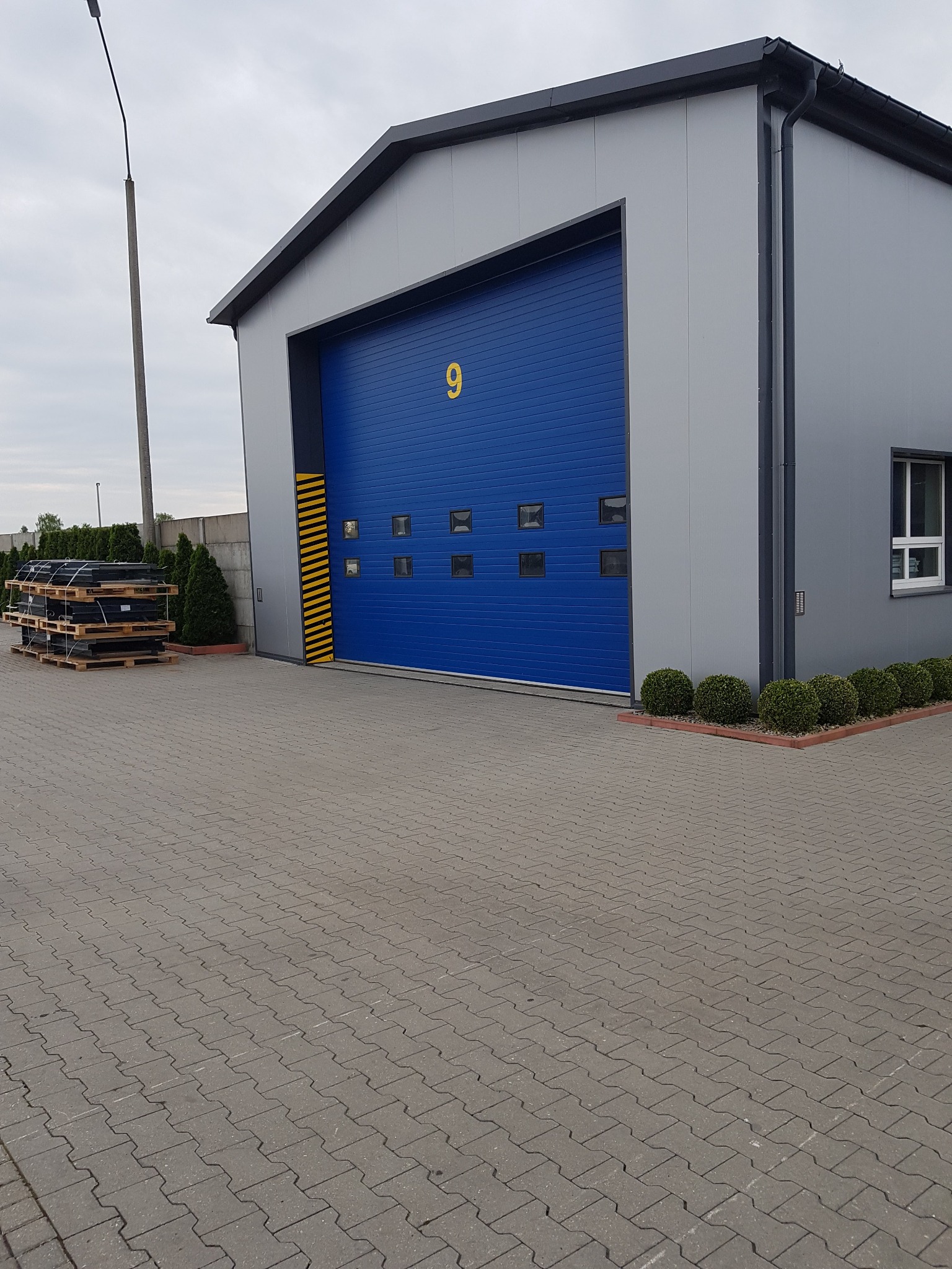 Gotowa hala z szarej blachy z niebieską bramą segmentową z numerem 9, widoczny fragment chodnika z kostki brukowej i paleta z materiałami budowlanymi.