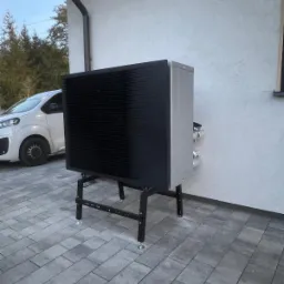 Zewnętrzna jednostka pompy ciepła powietrze-woda na czarnym stelażu, ustawiona na szarej kostce brukowej przy jasnej ścianie budynku. Widoczny fragment białego samochodu.