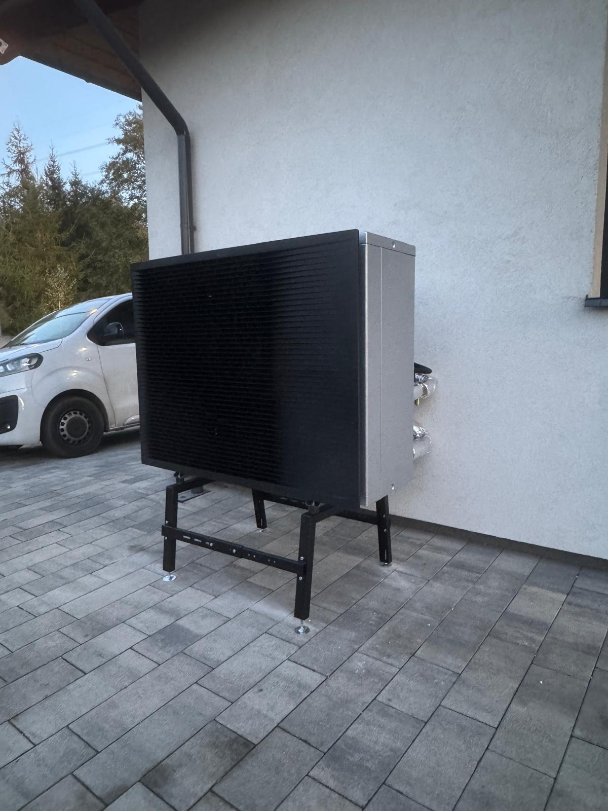 Zewnętrzna jednostka pompy ciepła powietrze-woda na czarnym stelażu, ustawiona na szarej kostce brukowej przy jasnej ścianie budynku. Widoczny fragment białego samochodu.