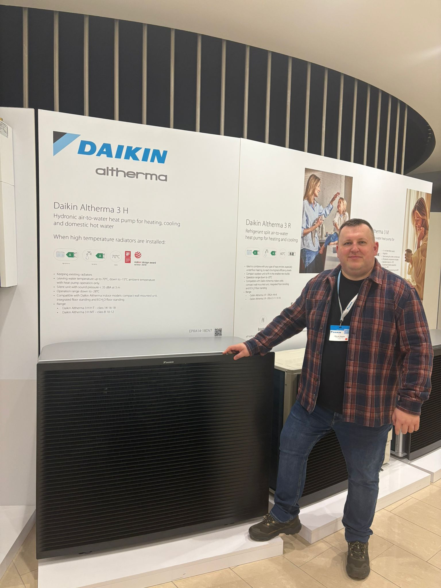 Prezentacja pompy ciepła Daikin Altherma 3H na stoisku targowym. Mężczyzna w koszuli w kratę stoi obok urządzenia. Widoczne logo Daikin i informacje o produkcie.