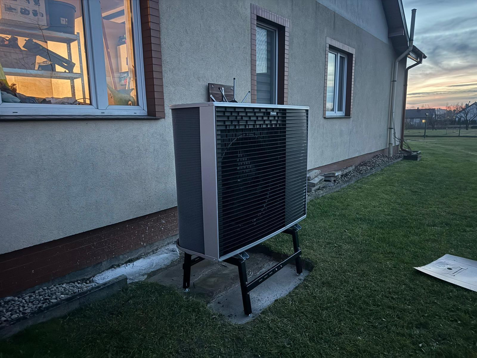 Pompa ciepła Daikin na zewnątrz budynku, zamontowana na czarnej konstrukcji wsporczej. Widoczne okna budynku i trawnik. Zachodzące słońce w tle.
