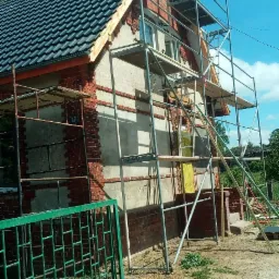 Dom w trakcie remontu z nowym ciemnym dachem, widoczne rusztowania i fragmenty odsłoniętej cegły na elewacji.