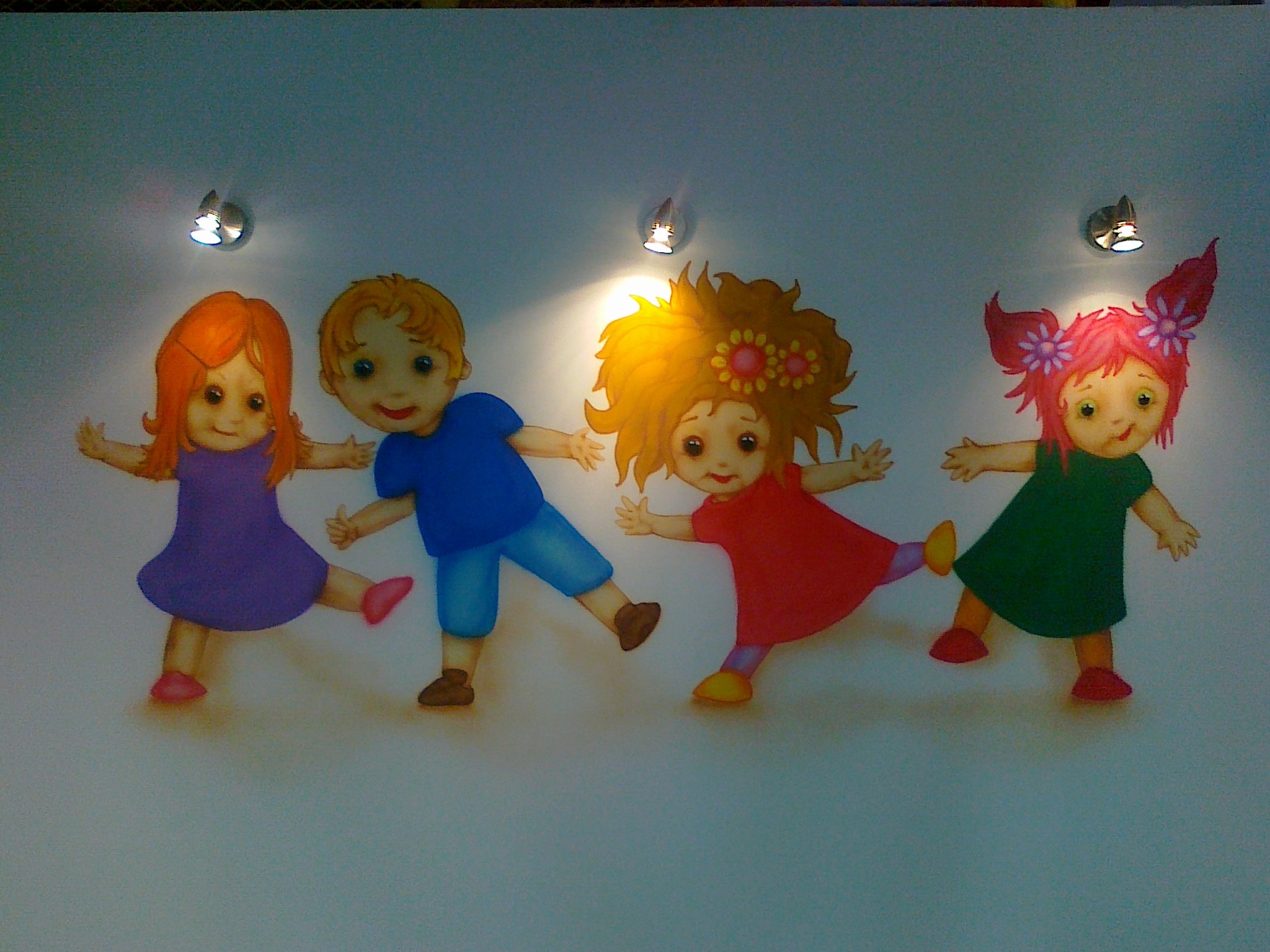 Mural z czterema postaciami dzieci w różnych kolorowych strojach, trzymających się za ręce, oświetlony trzema punktowymi lampami sufitowymi.