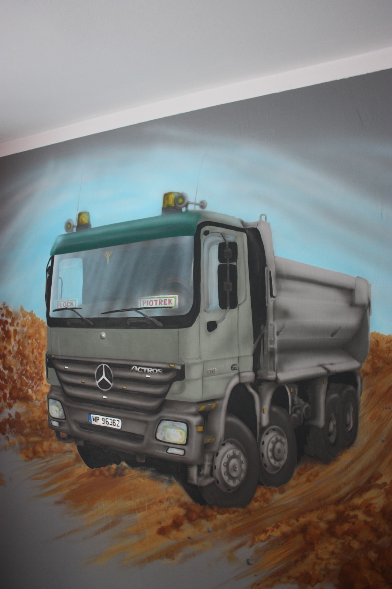 Iluzjonistyczne malowidło ścienne przedstawiające ciężarówkę Mercedes-Benz Actros w trakcie jazdy po terenie budowy, z widocznymi detalami jak tablice rejestracyjne i napisy na szybie.