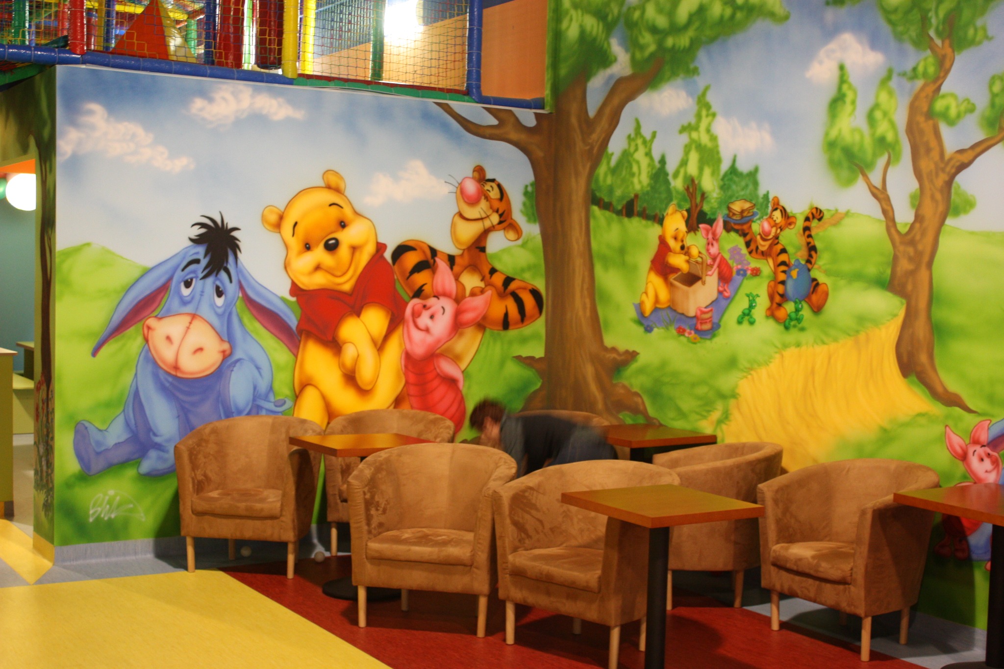 Wnętrze sali zabaw z malowidłem ściennym przedstawiającym postacie z Kubusia Puchatka, w tle widoczna konstrukcja zabawowa, a na pierwszym planie brązowe fotele i stoliki.