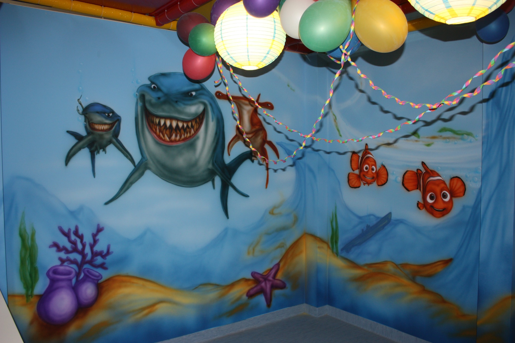 Ściana pokoju dziecięcego z ręcznie malowanym podwodnym światem: rekiny, błazenki, rozgwiazda, koralowce, stylizowane na bajkę 'Gdzie jest Nemo', dekorowana balonami i lampionami.